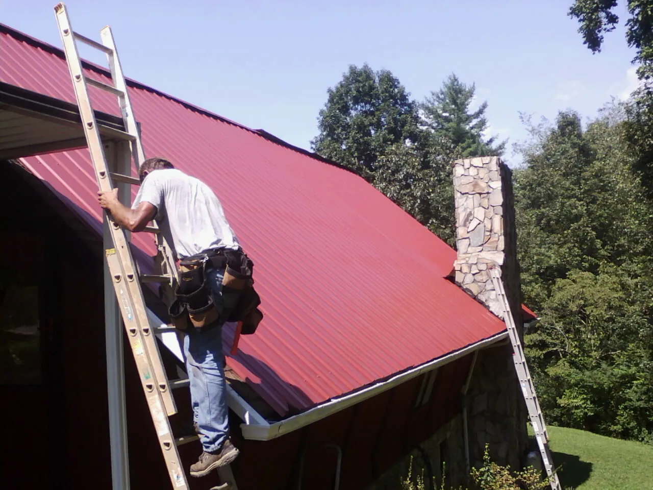 Roofer on a ladder beside a red metal roof and stone chimney — Soffit & Fascia Repair by Roofing Brothers in Gladstone