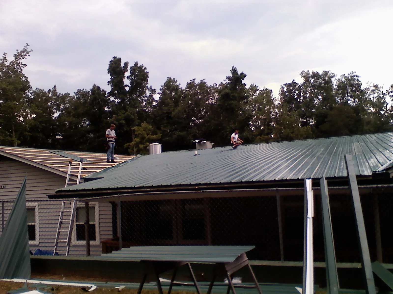 Two roofers installing green metal panels above a covered porch in Gladstone